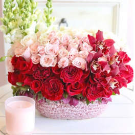 Pink and red rose arrangement in a round basket with a candle beside it.