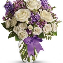 White roses and purple flowers arranged in a glass vase with a purple ribbon