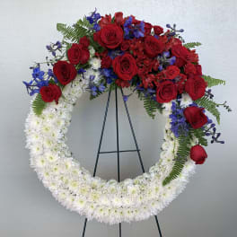 Red roses and blue flowers arranged in a large white floral wreath on a stand.