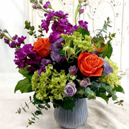 Vibrant arrangement of orange roses, purple stock, and green hydrangeas in a ribbed gray vase