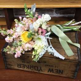 Loose hand-tied wedding bouquet of pastel blooms with a yellow rose and sage ribbon on a wooden crate.