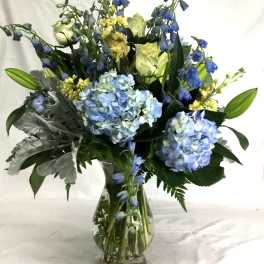 Tall arrangement of blue hydrangeas, white roses, lilies, and yellow flowers in a clear glass vase.
