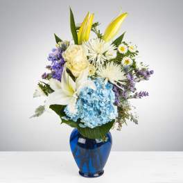 Mixed bouquet of blue hydrangea, white lilies, roses, and daisies in a blue glass vase