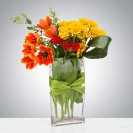 Tall glass vase of orange tulips, yellow daisies, and red roses with a green ribbon.