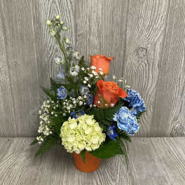 Mixed bouquet in an orange vase with orange roses, blue flowers, and pale hydrangea