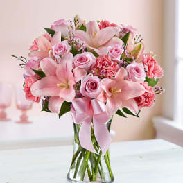 Pink lilies, roses, and carnations in a glass vase with a pink ribbon