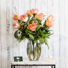 Peach and pink roses with small white blooms arranged in a clear glass vase on a rustic table