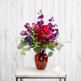 Red, pink, and purple mixed flower arrangement in a red glass vase on a small table with a candle