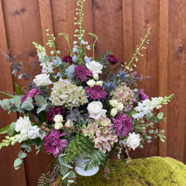 Large floral arrangement with white roses, purple mums, and pale hydrangeas in a white vase.
