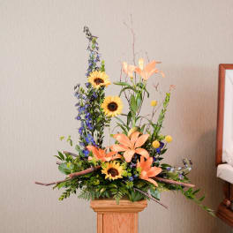 Tall floral arrangement with peach lilies, sunflowers, and blue delphinium in a wooden pedestal vase