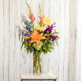 Tall mixed bouquet with orange lilies, yellow daisies, and purple blooms in a glass vase