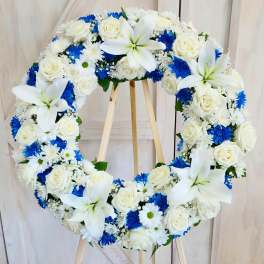Heart-shaped floral wreath with white roses, lilies, and blue daisies on a wooden stand