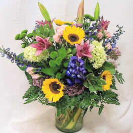 Mixed arrangement with sunflowers, lilies, roses, hydrangeas and delphinium in a glass vase