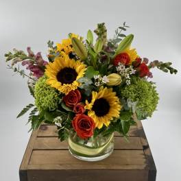 Arrangement of sunflowers, red roses, and mixed blooms in a clear glass vase on a wooden crate.