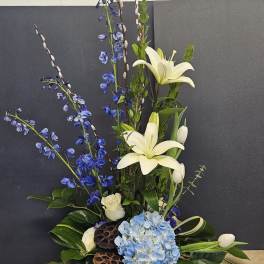 Tall floral arrangement with white lilies, blue flowers, and hydrangea in a low black base