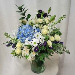 Mixed arrangement of blue hydrangea, white daisies and roses, and purple flowers in a clear glass vase