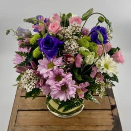 Round arrangement of pink roses, purple lisianthus, and pastel mums in a clear glass vase