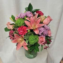 Mixed bouquet of pink lilies, roses, carnations, and lavender mums in a clear glass vase.