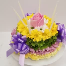 Round floral cake of yellow and pink blooms with a pink rose, purple bows, and tall white candles