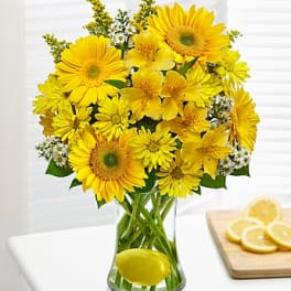 Bright yellow daisy and gerbera bouquet in a clear vase with a lemon, beside sliced lemons on a board