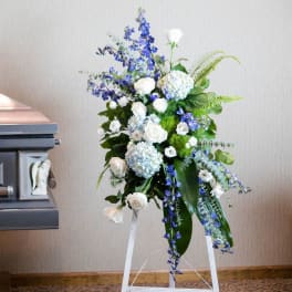Tall funeral spray with white roses, blue flowers, and hydrangeas on an easel