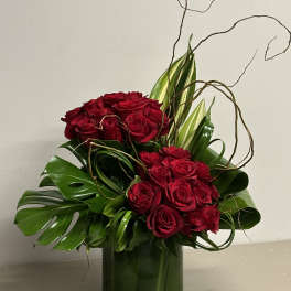 Tall arrangement of red roses with tropical green leaves and curly branches in a clear glass cylinder vase