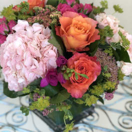 Compact arrangement of orange roses, pink hydrangeas, and hot pink spray roses in a glass cube vase