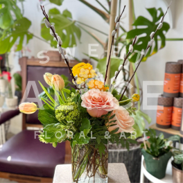 Peach roses, yellow blooms, and tulips in a clear glass vase with tall pussy willow branches.
