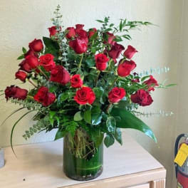Tall arrangement of red roses with lush foliage in a clear glass cylinder vase
