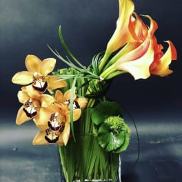Orange calla lilies and orchids arranged in a clear glass vase