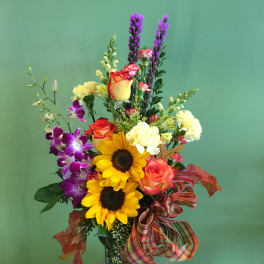 Mixed bouquet with sunflowers, roses, orchids, and purple spikes in a glass vase