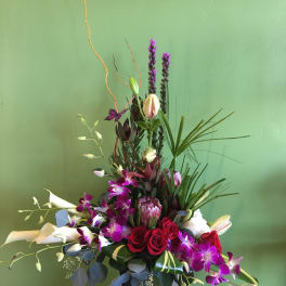 Tall floral arrangement with red roses, purple orchids, and white calla lilies in a white vase