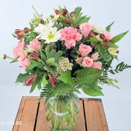 Mixed bouquet of pink roses, carnations, and white daisies in a clear glass vase on a wooden table.