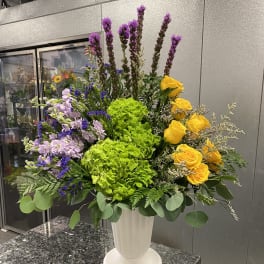 Tall floral arrangement with yellow roses, green hydrangeas, and purple flowers in a white vase
