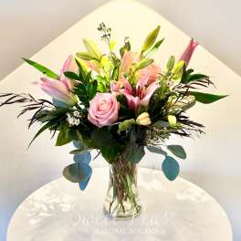 Pink lilies and roses with white accent blooms arranged in a clear glass vase.