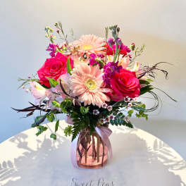 Pink roses and pale gerbera daisies arranged with mixed blooms in a pink glass vase with ribbon.