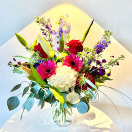 Bouquet of red roses, pink gerbera daisies, white hydrangea, and purple blooms in a glass vase