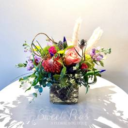 Mixed floral arrangement in a square glass vase with a pink rose and purple blooms