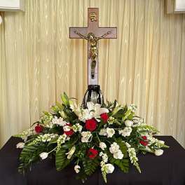 Sympathy floral arrangement with red roses, white lilies, and a crucifix backdrop