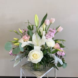 Pastel arrangement of white lilies and roses with pink tulips and stock in a glass cube vase.