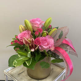 Pink roses and lilies arranged in a gold vase with a pink ribbon