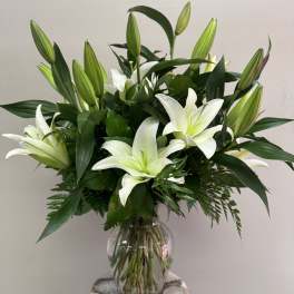 Tall arrangement of white lilies with green buds in a clear glass vase.