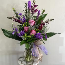Tall arrangement of pink roses and purple flowers in a clear glass vase with decorative bow