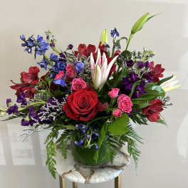 Colorful mixed arrangement with red roses, pink lilies, and purple blooms in a clear glass vase