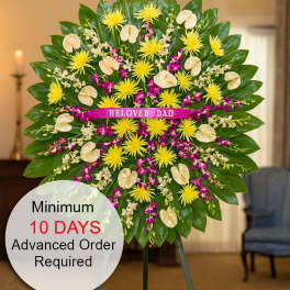 Tall standing sympathy spray with yellow mums, cream tropical blooms and purple orchids on a green leaf fan.