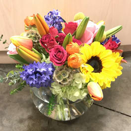 Colorful mixed bouquet in a clear glass vase with sunflowers, roses, tulips, and purple blooms