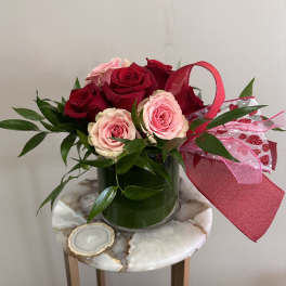 Short arrangement of red and pink roses with greenery in a glass vase, accented with a red and pink bow.