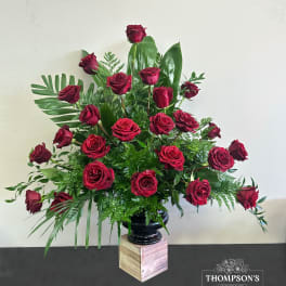 Tall arrangement of red roses with lush greenery in a black vase on a wooden pedestal.