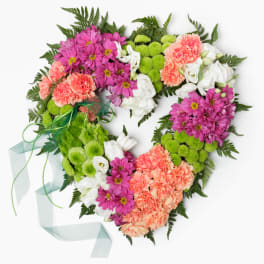 Heart-shaped wreath of pink mums, peach carnations, green button poms, and white blooms with green ribbon.
