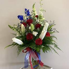 Red roses and white lilies in a clear glass vase with red, white, and blue ribbons
